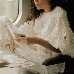 May include: A woman wearing a white, lacy dress with a floral pattern sits on a train and holds a deck of cards in her hands.