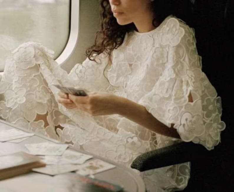 May include: A woman wearing a white, lacy dress with a floral pattern sits on a train and holds a deck of cards in her hands.