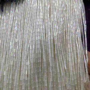 Pode incluir: Close-up de um feixe de contas de sementes brancas e iridescentes. As contas são longas e cilíndricas, penduradas em fios verticais. Uma pequena seção de contas azuis escuras e iridescentes é visível no topo. As contas são provavelmente usadas para fazer joias ou enfeites.