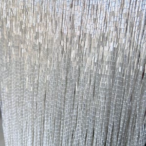 May include: Close-up of a curtain of silver-colored beads. The beads are small, cylindrical, and strung together in vertical strands. The light reflects off the beads, creating a shimmering effect. The overall effect is elegant and decorative.