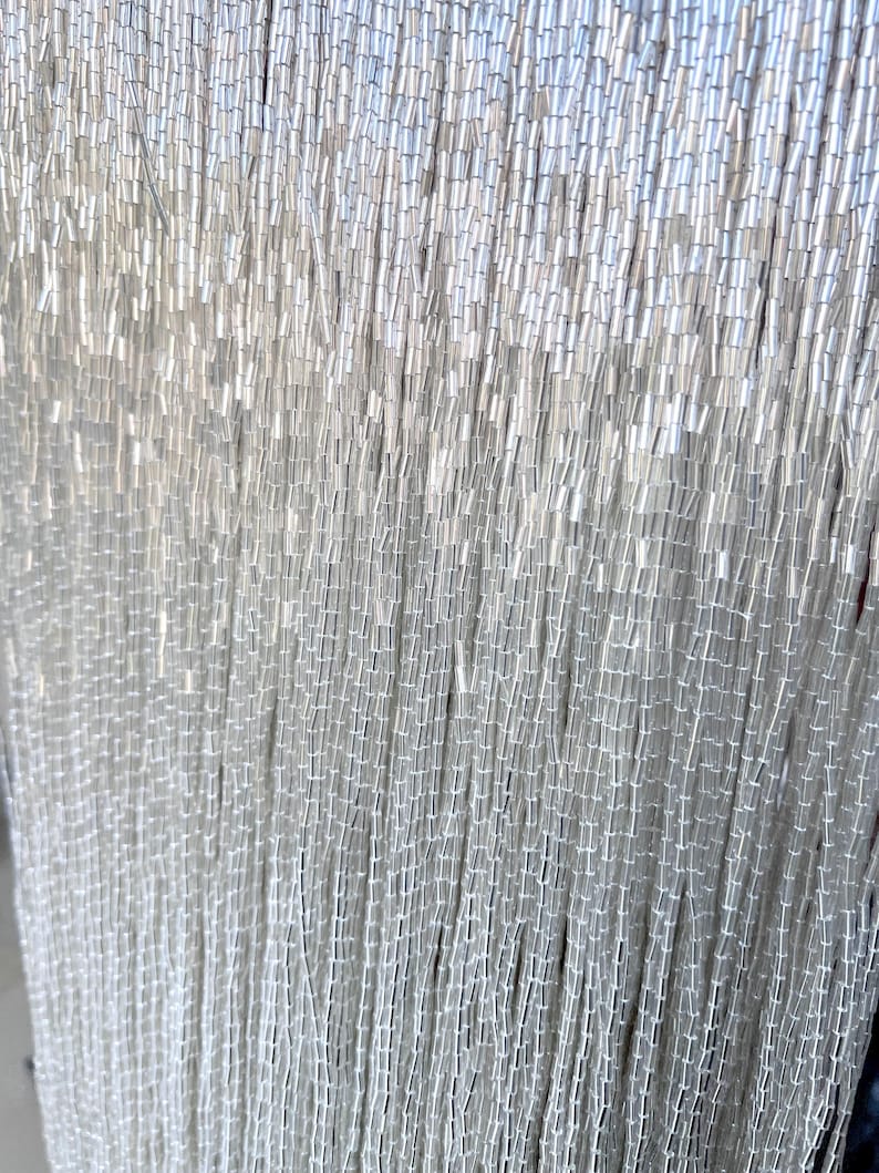 May include: Close-up of a curtain of silver-colored beads. The beads are small, cylindrical, and strung together in vertical strands. The light reflects off the beads, creating a shimmering effect. The overall effect is elegant and decorative.