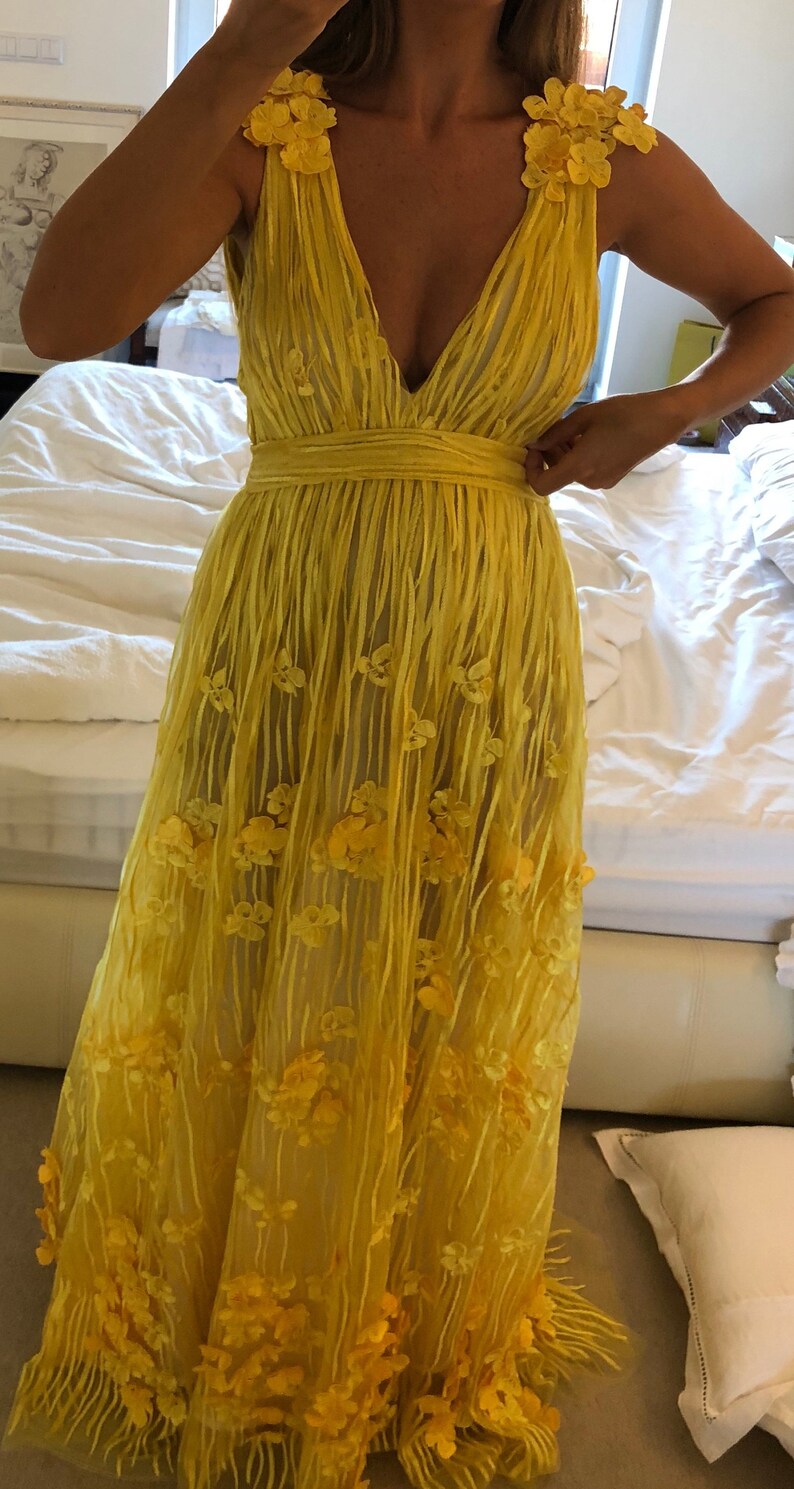 May include: A yellow maxi dress with a V-neckline and floral embellishments. The dress has a fitted bodice and a flowing skirt.