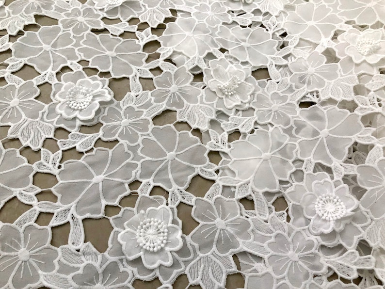 May include: White lace fabric with a floral design. The fabric has a delicate, intricate pattern with raised flowers.