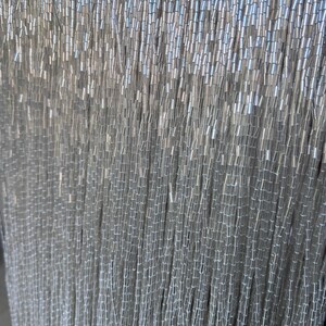 May include: Close-up of a curtain of silver beads. The beads are small, cylindrical, and strung together in vertical strands. The overall effect is a shimmering, textured surface, ideal for decorative purposes.