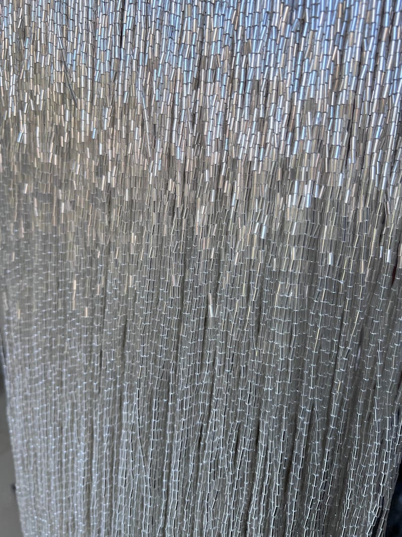 May include: Close-up of a curtain of silver beads. The beads are small, cylindrical, and strung together in vertical strands. The overall effect is a shimmering, textured surface, ideal for decorative purposes.