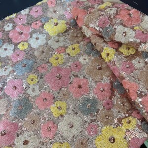 Könnte beinhalten: Ein Stoff mit floralem Muster. Der Stoff ist mit Pailletten bedeckt und weist dreidimensionale Blüten in den Farbtönen Rosa, Gelb, Weiß, Grau und Braun auf. Die Blüten sind über die Oberfläche des Stoffes verstreut.