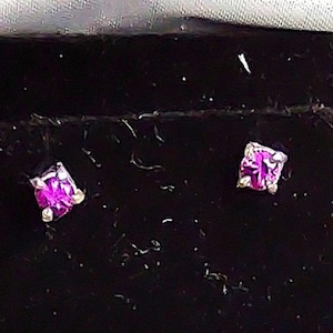 May include: A pair of square-cut, magenta gemstone stud earrings. The earrings are set in a silver metal, presented on a black velvet surface inside a jewelry box. The earrings are small and delicate.
