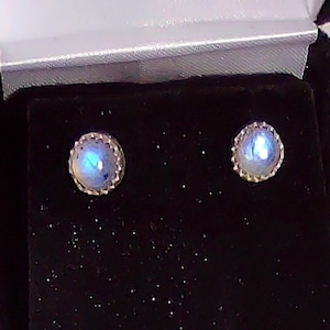 May include: A pair of moonstone stud earrings. Each earring features a round, iridescent moonstone set in a silver-colored, scalloped bezel. The earrings are displayed in a black velvet jewelry box with a white satin lining.
