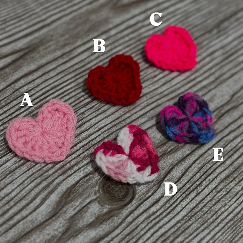 May include: Five crocheted hearts in different colours and patterns, including pink, red, hot pink, white, and purple. The hearts are arranged on a wooden surface.