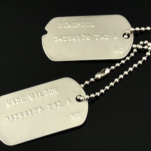 Puede incluir: Dos chapas de identificaci&oacute;n militares de plata en una cadena de plata. La chapa superior dice "DEADPOOL 34256178 T42 A" y la chapa inferior dice "WADE WILSON 34256178 T42 A".