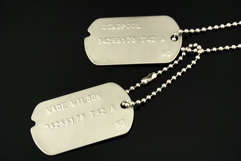 Puede incluir: Dos chapas de identificaci&oacute;n militares de plata en una cadena de plata. La chapa superior dice "DEADPOOL 34256178 T42 A" y la chapa inferior dice "WADE WILSON 34256178 T42 A".