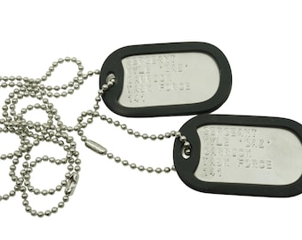 May include: Two military-style dog tags with ball chain necklaces. The tags are silver-colored with black rubber borders. Each tag has engraved text, including the name "Kyle 'SAZ' Carrick" and "Task Force 141".