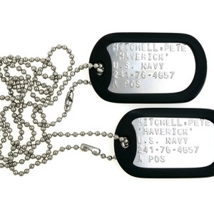 May include: Two black dog tags with silver lettering on a silver chain. The dog tags read "MITCHELL PETE MAVERICK U.S. NAVY 241-76-4657 A POS".