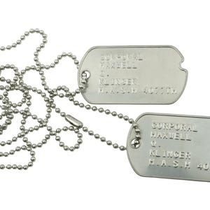 May include: Two silver dog tags on ball chains. Each tag is rectangular with rounded edges and features engraved text. The text includes the name "Maxwell" and "Klinger", along with other details. The chains are draped across each other.