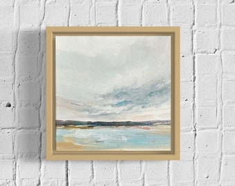May include: Square framed abstract landscape painting. The artwork features a serene scene with a light blue and tan body of water, a dark horizon line, and a cloudy sky. The frame is a light beige color.