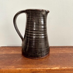 May include: A handmade, dark brown ceramic pitcher with a curved handle and a spout. The pitcher has horizontal textured lines around the body and sits on a wooden surface. The pitcher is approximately 7 inches tall.