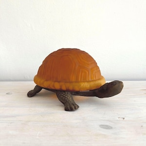 May include: A decorative turtle figurine with an amber-colored shell and dark green legs and head. The shell has a textured, geometric pattern. The figurine is displayed on a light-colored wooden surface.