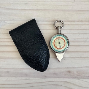 May include: A vintage compass with a black leather case. The compass has a white, red, and blue face with the words "Compass & Company." The case is textured and has a flap closure. The compass is a navigational tool.
