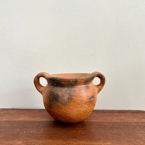 May include: A small, round, terracotta-colored ceramic pot with two loop handles. The pot has dark brown and black markings. The pot is sitting on a wooden surface.