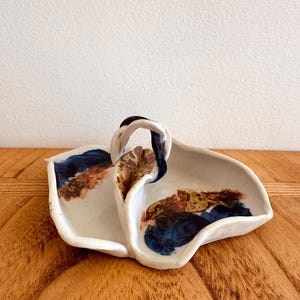 May include: A white ceramic serving dish with a handle. The dish has an abstract design in shades of blue, brown, and gold. The handle is black and white. The dish is on a wooden surface.