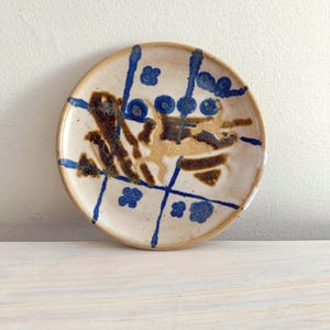 May include: A small, round ceramic plate with a cream-colored base. The plate is decorated with blue and brown abstract designs, including lines, dots, and floral shapes. The rim is a darker tan color. The plate is sitting on a light-colored wooden surface.