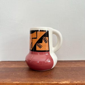May include: A small, white ceramic mug with a rounded base and a handle. The mug features a geometric design in orange, black, and white. The bottom half is a deep red color.