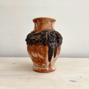 May include: A handmade ceramic vase with a textured, dark brown band around the middle. The vase has a light brown rim and base, with dark brown drips down the side. The vase sits on a light-colored wooden surface.