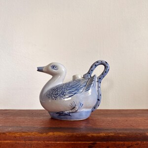 May include: A ceramic teapot shaped like a swan, with a white body and blue wing and tail details. The handle is a curved blue design. The spout is a small beak. The teapot sits on a blue base.