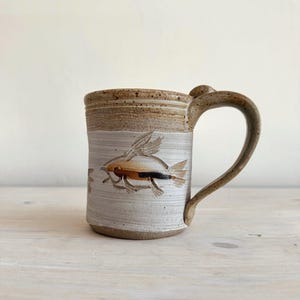 May include: A white ceramic mug with a black handle and the text "But First, Coffee" in a bold, black font. The mug is set against a blurred background, suggesting a cozy setting. The mug appears to be approximately 4 inches tall.