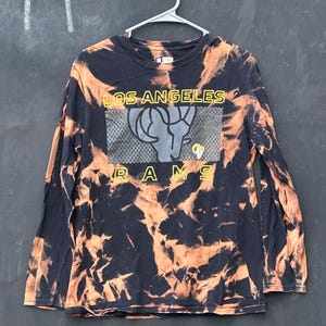 May include: Black long-sleeve t-shirt with a tie-dye pattern in orange and black. The shirt features a graphic of a ram's head with the text "LOS ANGELES RAMS" in yellow.