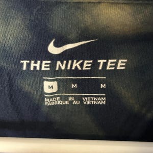 Puede incluir: Una camiseta azul tie-dye con un logotipo blanco de Nike y el texto "THE NIKE TEE". La etiqueta dice "M" y "MADE IN VIETNAM FABRIQUE AU VIETNAM".