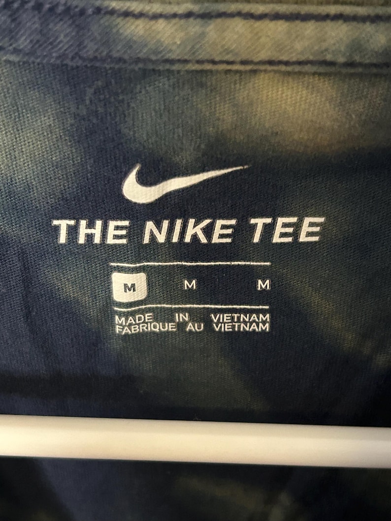 Puede incluir: Una camiseta azul tie-dye con un logotipo blanco de Nike y el texto "THE NIKE TEE". La etiqueta dice "M" y "MADE IN VIETNAM FABRIQUE AU VIETNAM".