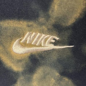 Puede incluir: Un logotipo Nike swoosh blanco bordado en una tela de tejido te&ntilde;ido anudado verde y amarillo.