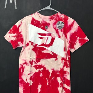 Könnte beinhalten: Rot-weißes Batik-T-Shirt mit einem weißen Logo eines stilisierten Buchstabens "D" und dem Text "Stadium Series" auf der Vorderseite. Das Shirt hängt an einem Kleiderbügel.