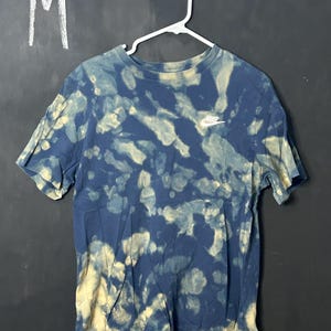Puede incluir: Una camiseta azul y blanca tie-dye con un logotipo blanco de Nike en la parte delantera. La camiseta est&aacute; colgada de una percha blanca. La talla "M" est&aacute; escrita sobre un fondo negro.
