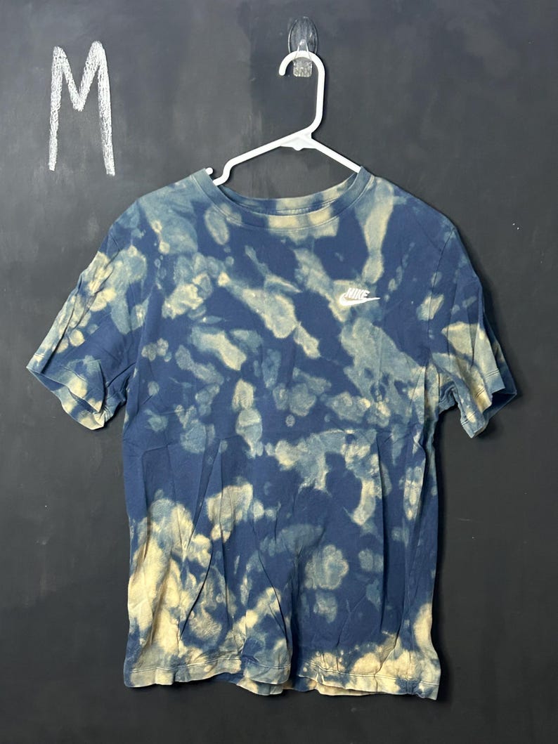 Puede incluir: Una camiseta azul y blanca tie-dye con un logotipo blanco de Nike en la parte delantera. La camiseta est&aacute; colgada de una percha blanca. La talla "M" est&aacute; escrita sobre un fondo negro.
