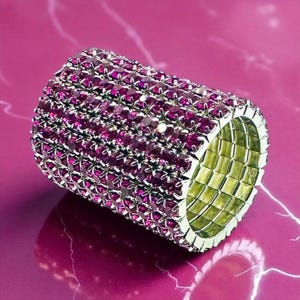 May include: A wide, cylindrical ring covered in small, vibrant pink gemstones. The ring has a silver-colored metal band and a grid-like pattern on the inside. The ring is set against a pink background.