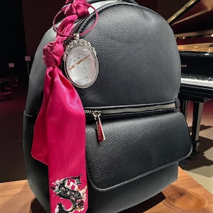 May include: Black backpack with a front pocket and gold zipper. A pink scarf with a floral design and the letter "S" is attached to the zipper. A silver pendant with a white face hangs from the top.