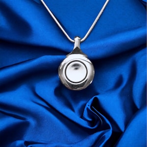 May include: A silver pendant necklace with a circular design. The pendant is suspended from a thin, silver chain. The necklace is displayed on a vibrant blue satin fabric, creating a striking contrast.