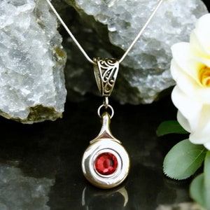 May include: A silver necklace with a pendant featuring a round, silver charm with a red gemstone in the center. The pendant has a delicate, ornate design.