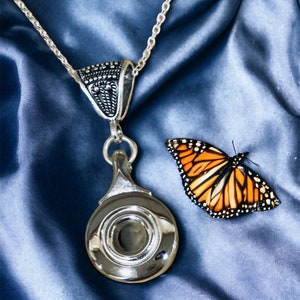 May include: A silver necklace with a detailed pendant shaped like a magnifying glass. The pendant has a round, clear glass lens. The necklace is on a blue satin background with a butterfly.
