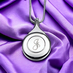 May include: A silver pendant necklace with a round charm featuring the letter "J" in a cursive script. The pendant is on a silver chain and is displayed on a purple fabric background.