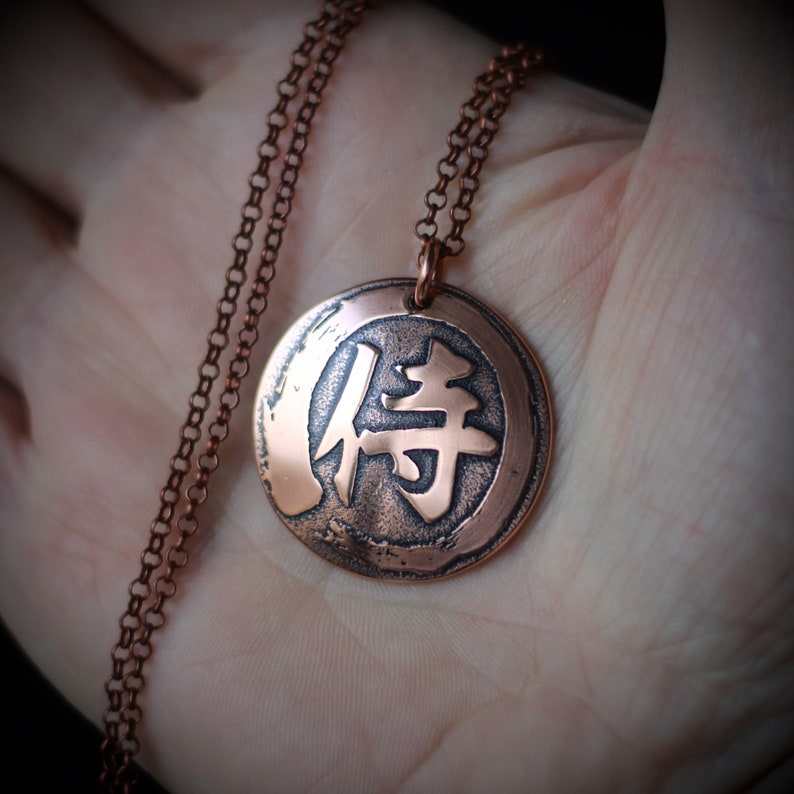 May include: Copper-colored pendant necklace featuring a circular pendant with a Japanese Kanji symbol. The pendant hangs from a matching chain. The Kanji symbol is in a darker tone, creating contrast against the copper background. The necklace is displayed on a hand.
