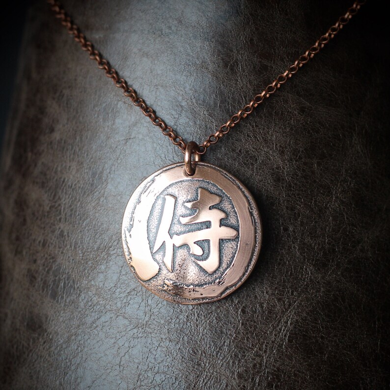 May include: A copper-colored pendant necklace featuring a circular charm with a Japanese character. The charm and chain have a rustic finish. The necklace is displayed against a textured, dark background.