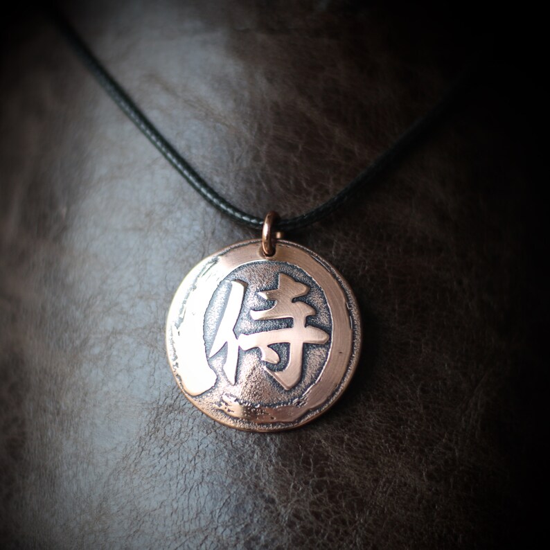 May include: Copper-colored round pendant necklace with a black cord. The pendant features a Japanese character in the center. The pendant is approximately 1.5 inches in diameter. The necklace is displayed against a brown leather background.