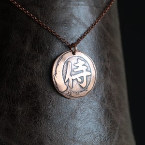 May include: Copper-colored pendant necklace featuring a circular pendant with a Japanese Kanji symbol. The pendant hangs from a matching copper chain. The pendant has a textured, hammered appearance. The background is a dark brown leather-like material.