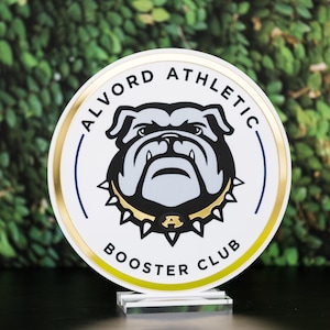May include: A round, white sign with the text "ALVORD ATHLETIC BOOSTER CLUB" in black lettering. The sign features a bulldog graphic in black, white, and gray, with a gold-colored trim and a clear acrylic base.