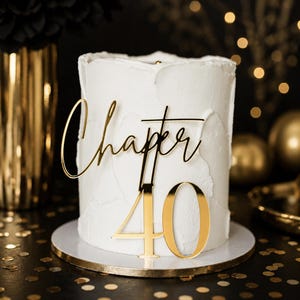 May include: A white cylindrical cake with textured frosting features a gold-colored acrylic cake topper. The topper displays the text "Chapter 40" in a combination of cursive script for the word "Chapter" and bold block numerals for "40".