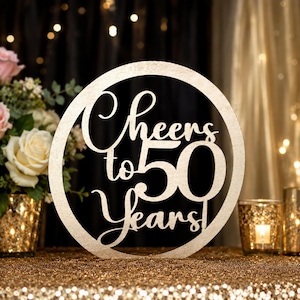 May include: A wooden circular sign with the words "Cheers to 50 Years!" cut out in a decorative font. The sign is light-colored and set against a background of gold and black, with flowers and candles in the background.