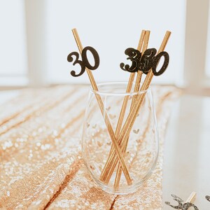 May include: Gold straws with black glitter numbers 30 on them in a clear glass with a heart pattern. The straws are on a gold sparkly surface.